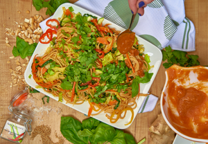 A shallow white plate filled with noodles and fresh ingredients such as basil, ginger, bell pepper, cilantro, zucchini, peanuts, sesame; plus a bowl of sauce and a hand spoons sauce over the dish