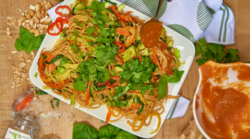 A shallow white plate filled with noodles and fresh ingredients such as basil, ginger, bell pepper, cilantro, zucchini, peanuts, sesame; plus a bowl of sauce and a hand spoons sauce over the dish